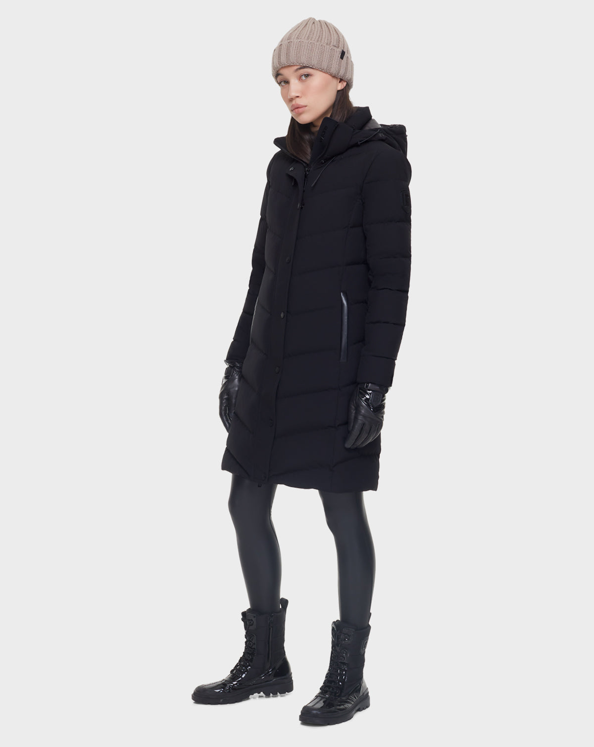 A person wears the Rudsak ELISA - L121512 BLACK womens quilted down puffer with black gloves, leggings, boots, and a beige knit beanie, standing against a plain white background.