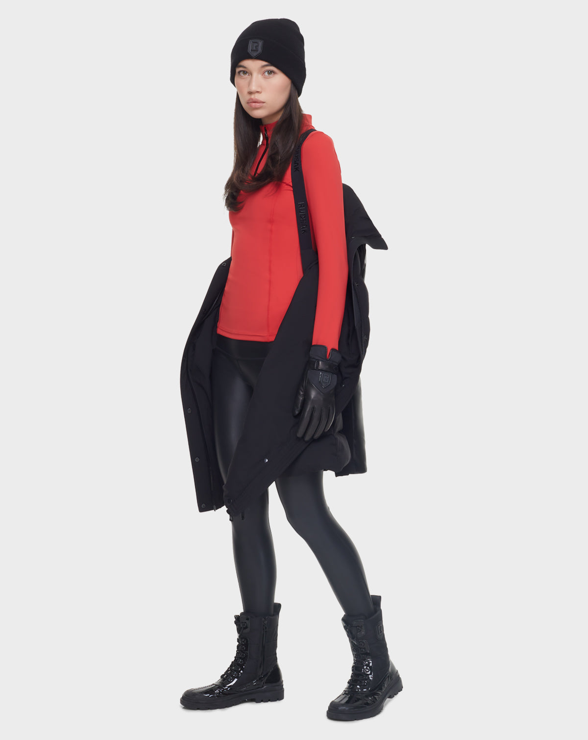 A woman wears the Rudsak ELSA - L121519 BLACK sustainable winter coat draped over her shoulders, paired with a black beanie, red fitted top, black gloves, shiny black leggings, and combat boots against a plain white background.