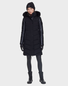 A woman faces forward wearing the Rudsak ELSA - L121519 BLACK sustainable winter coat with a fur-trimmed hood, black leather sleeves, leggings, lace-up boots, gloves, and a black beanie against a plain light background.