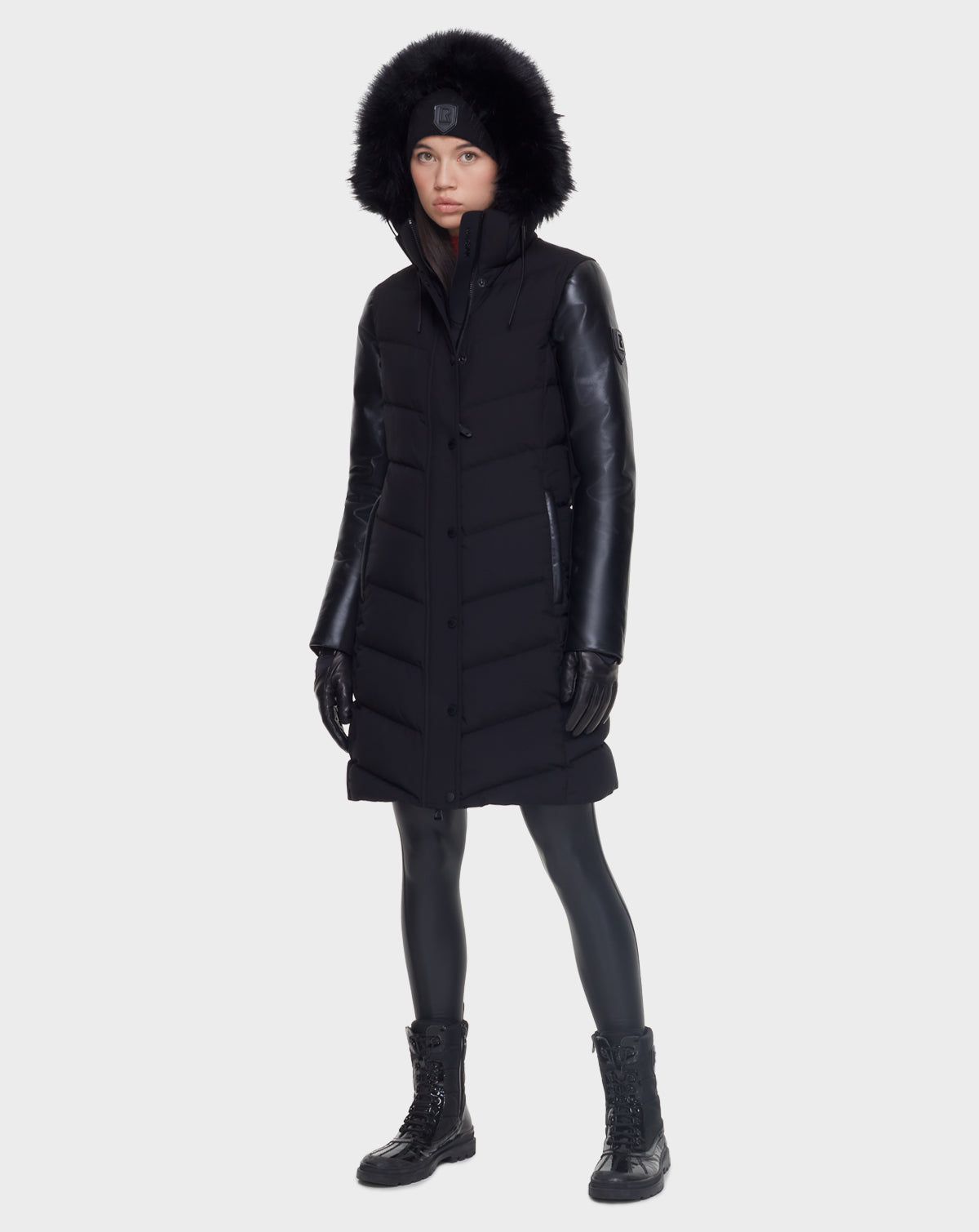 A person wearing the Rudsak ELSA - L121519 BLACK sustainable winter coat with a fur-trimmed hood, black leggings, lace-up black boots, gloves, and a beanie stands against a plain light background.