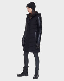 A person stands in profile wearing the Rudsak ELSA - L121519 BLACK women’s quilted down puffer with leather sleeves, black gloves, beanie, leggings, and lace-up boots—all set against a plain light background.