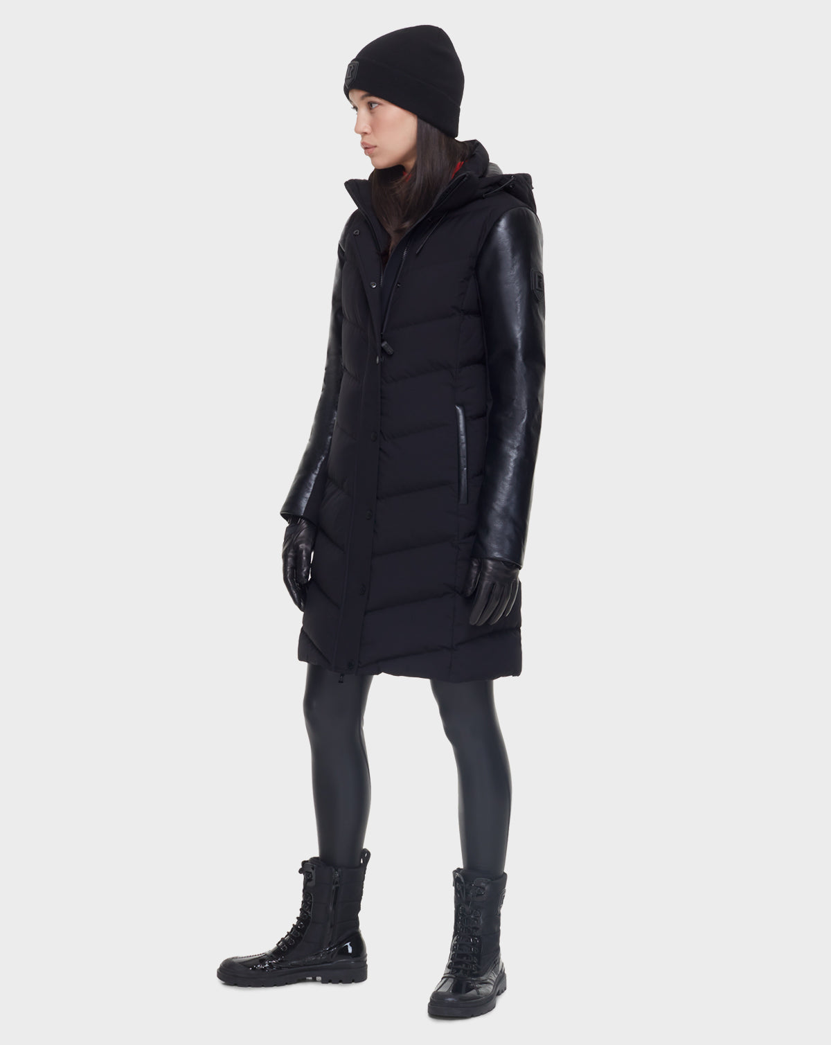A person stands in profile wearing the Rudsak ELSA - L121519 BLACK women’s quilted down puffer with leather sleeves, black gloves, beanie, leggings, and lace-up boots—all set against a plain light background.