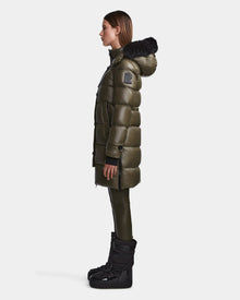 A woman stands in profile wearing the Rudsak MEYA S - L123521 Light Olive women's down puffer with a black fur-trimmed hood, paired with black pants and tall black boots against a plain white background.