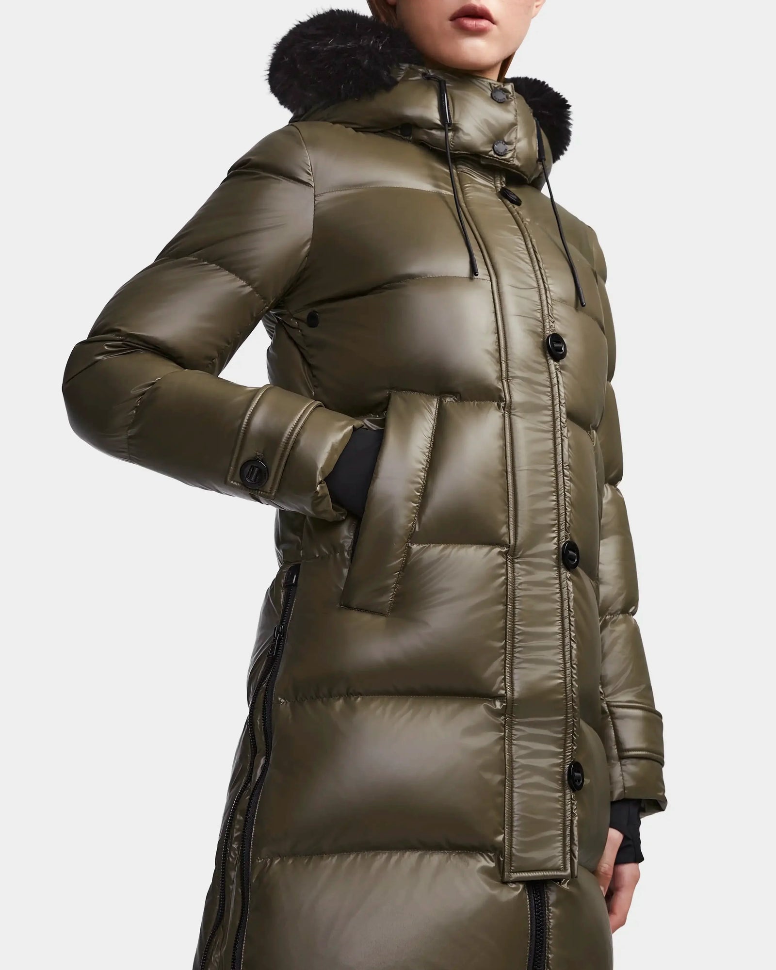 A person wears the Rudsak MEYA S - L123521 Light Olive women's down puffer coat with black buttons, a hood with dark fur trim, and hands in side pockets. The shiny olive green coat image is cropped at the nose and mid-thigh.