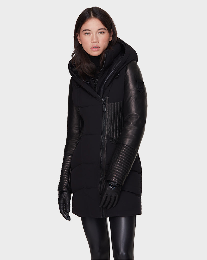 A woman stands facing forward against a plain light background wearing the Rudsak SHAUNA - 8121509 BLACK winter coat with leather sleeves, ribbed leather panels, and black gloves.