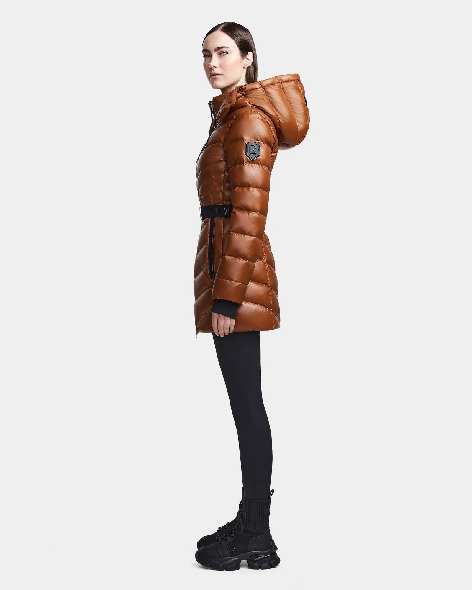 A woman stands in profile against a plain light background wearing the Rudsak MALAYA - L123510 Dark Camel shiny women’s down puffer jacket with a belt, made from recycled material, paired with black leggings and chunky boots.