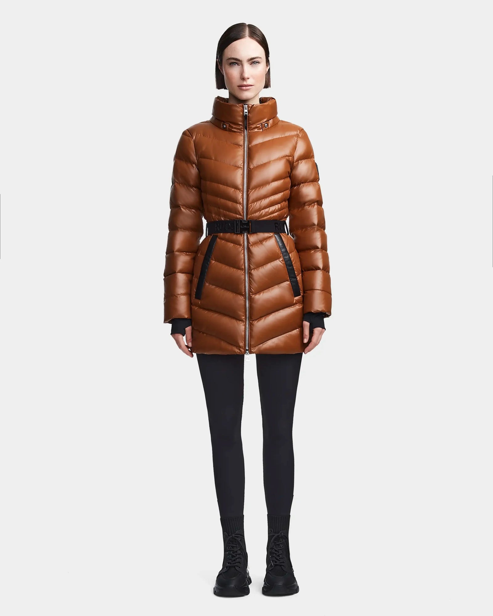 A woman wears the Rudsak MALAYA - L123510 Dark Camel shiny brown down puffer jacket with herringbone quilting, styled with black leggings and shoes, standing facing forward against a plain white background.