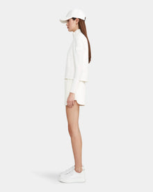 A woman stands in profile against a plain light background wearing the Rudsak LOUISE - 8124046 Eggshell womens hybrid knit sweater jacket, paired with a white cap, white shorts, and white platform sneakers.