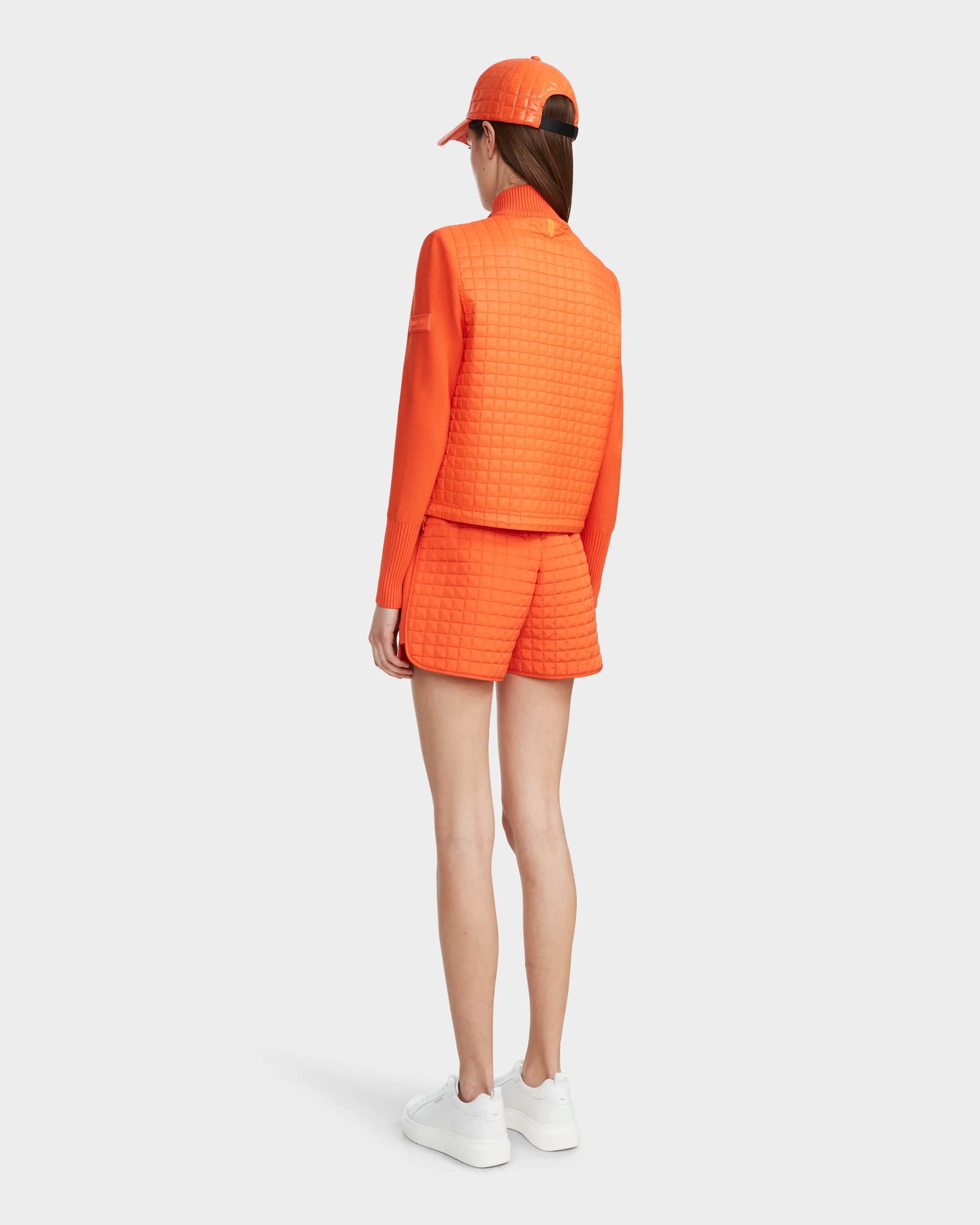 A person faces away, wearing the Rudsak LOUISE - 8124046 Orange Sorbet lightweight quilted jacket with matching shorts and a baseball cap, paired with white sneakers, against a plain white background.