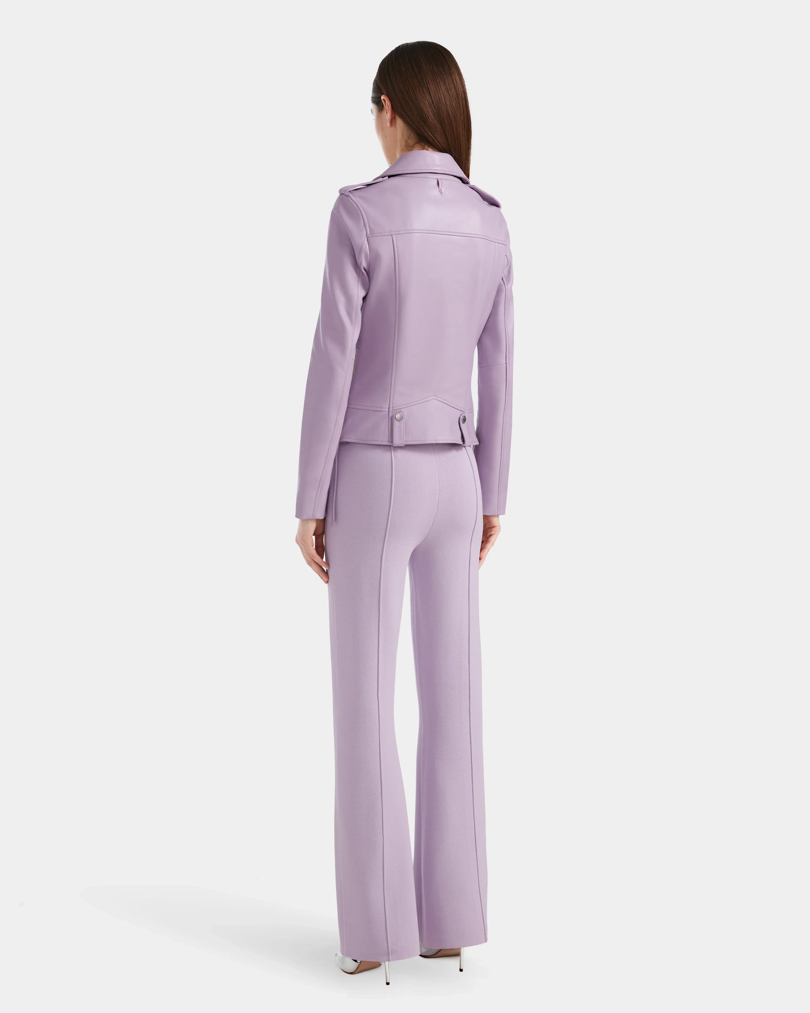 A woman with straight brown hair stands facing away, wearing the Rudsak MERGO - 8124034 Lilac Foam biker jacket and matching wide-legged pants with high heels against a plain white background.