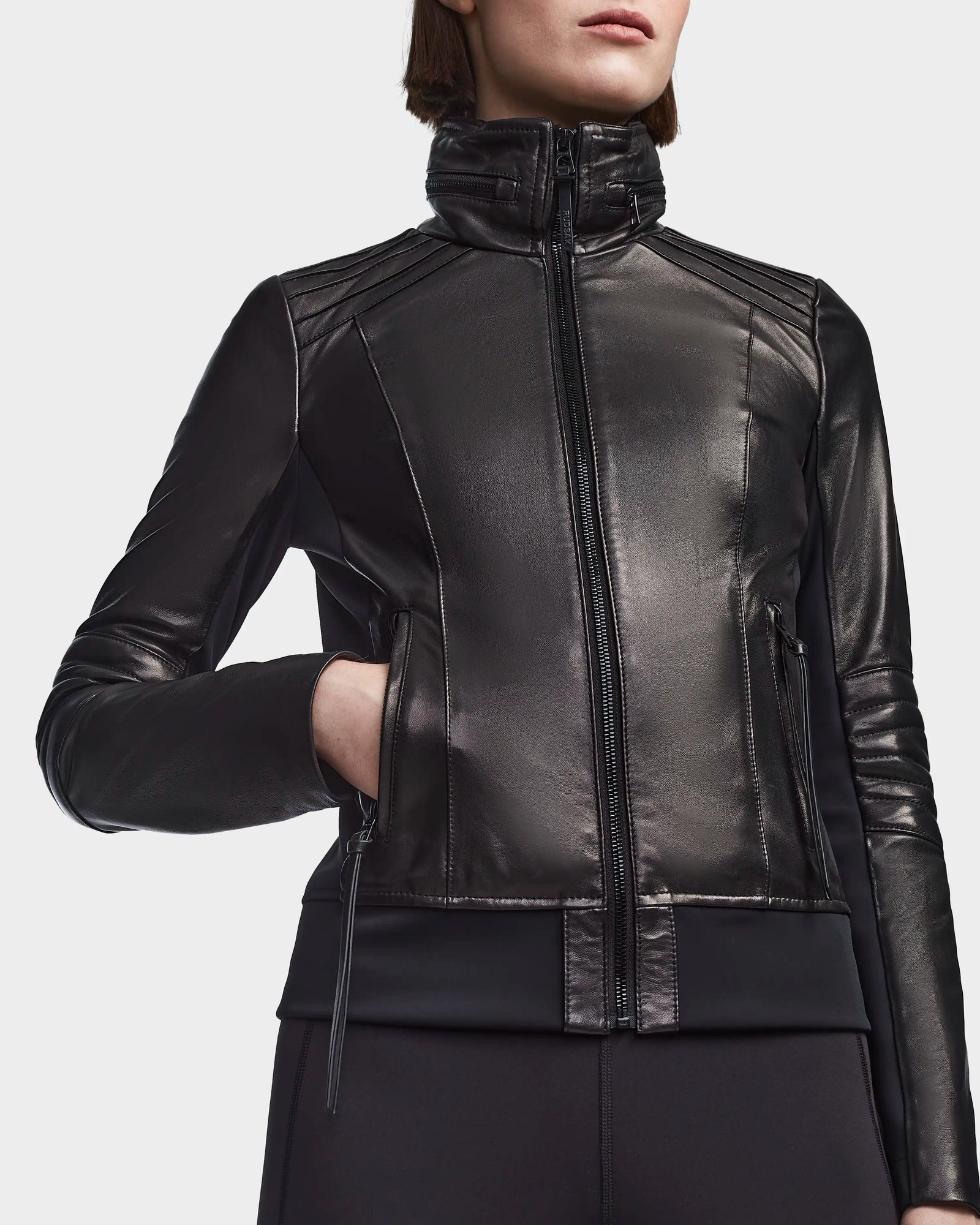 A person wears the Rudsak PRESLEY - 8122027 BLACK, a shiny black moto-style bomber jacket with a high collar and zipped pockets, standing with one hand in a pocket against a plain white background.