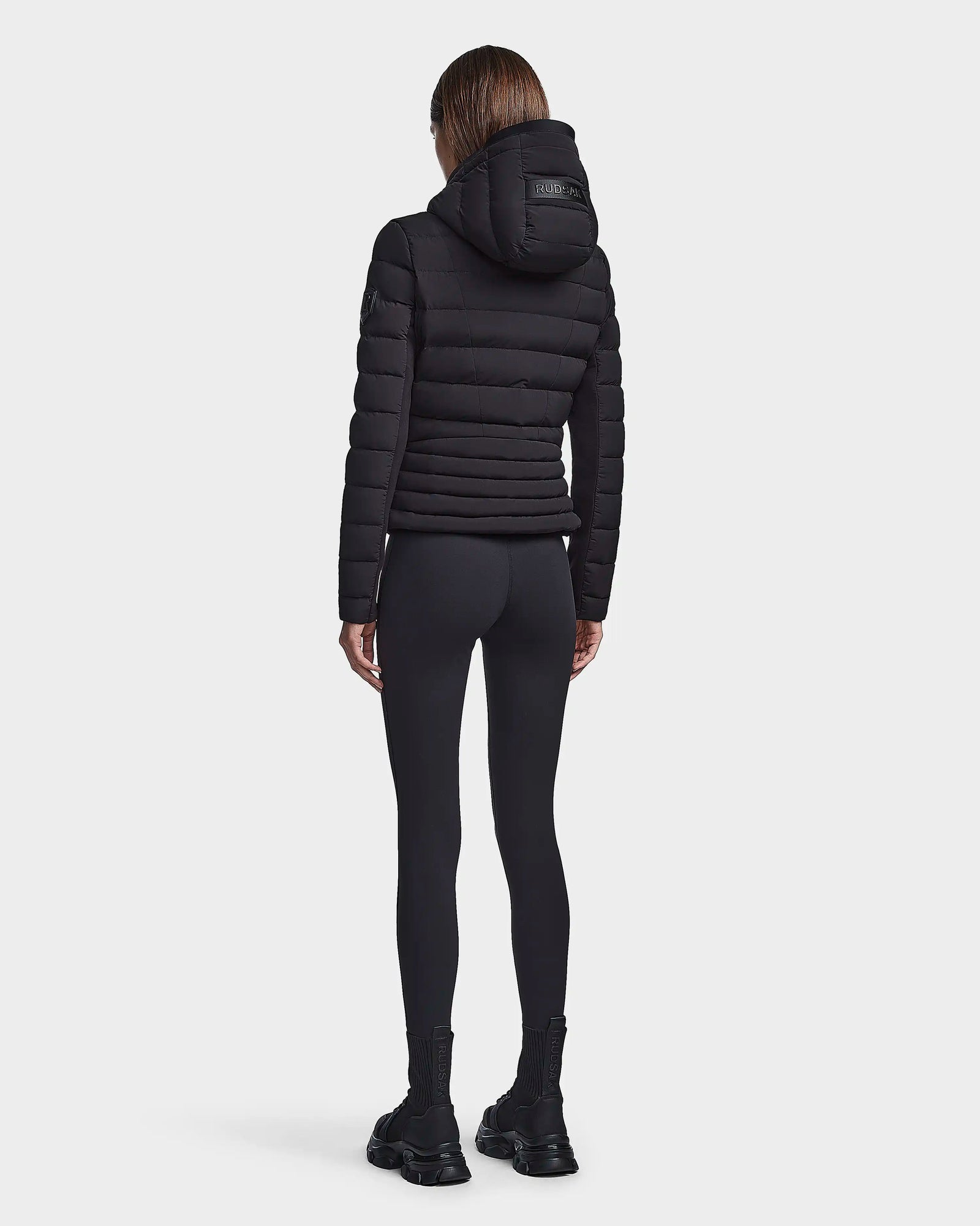 A person with straight brown hair, seen from behind, wears the RUDSAK MELISSA - 8122551 BLACK womens puffer jacket, black leggings, and black boots against a plain white background.
