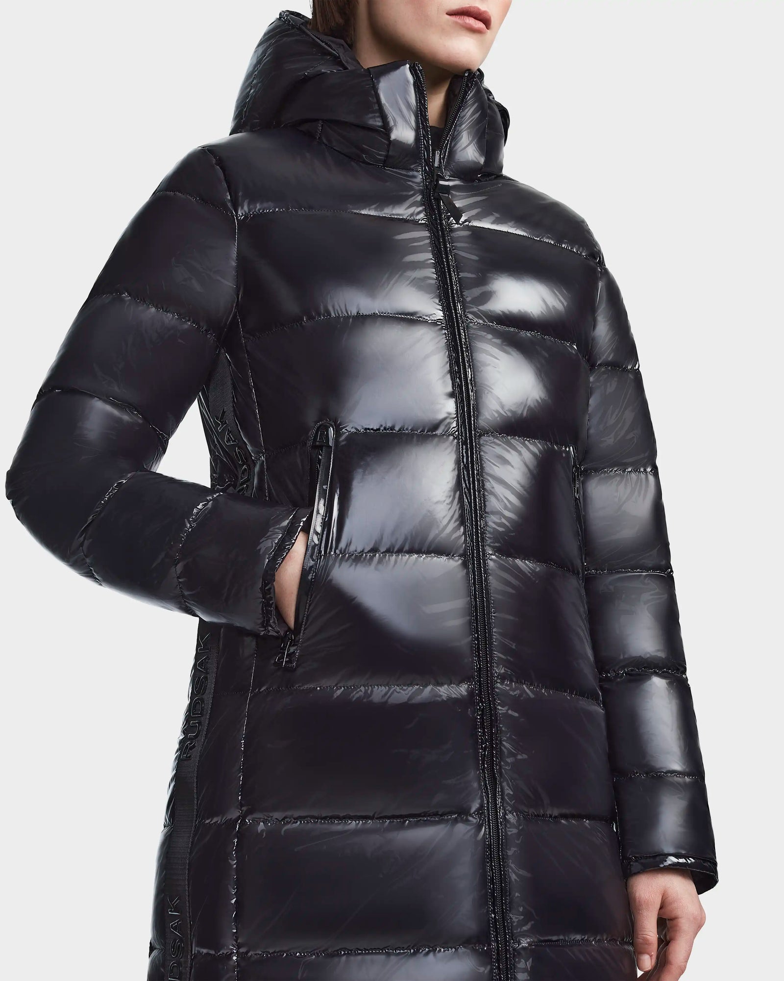 A person wears the RUDSAK MARNIE LONG - 8122519 BLACK womens puffer jacket with a hood and zippered front, one hand in the side pocket. The image is cropped to show their upper body and part of their face.