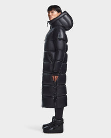 A woman stands in profile wearing the Rudsak RITA L-S - L123526 Black maxi down puffer with a large hood, quilted design, and R logo patch on the upper sleeve. She pairs it with platform boots against a plain light background.