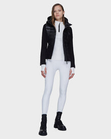 A woman stands against a plain background wearing Rudsak’s MABS - 8121012 BLACK lightweight activewear jacket with waterproof zippers, styled with a white zip-up top, white leggings, and black lace-up boots, her hands relaxed by her sides.
