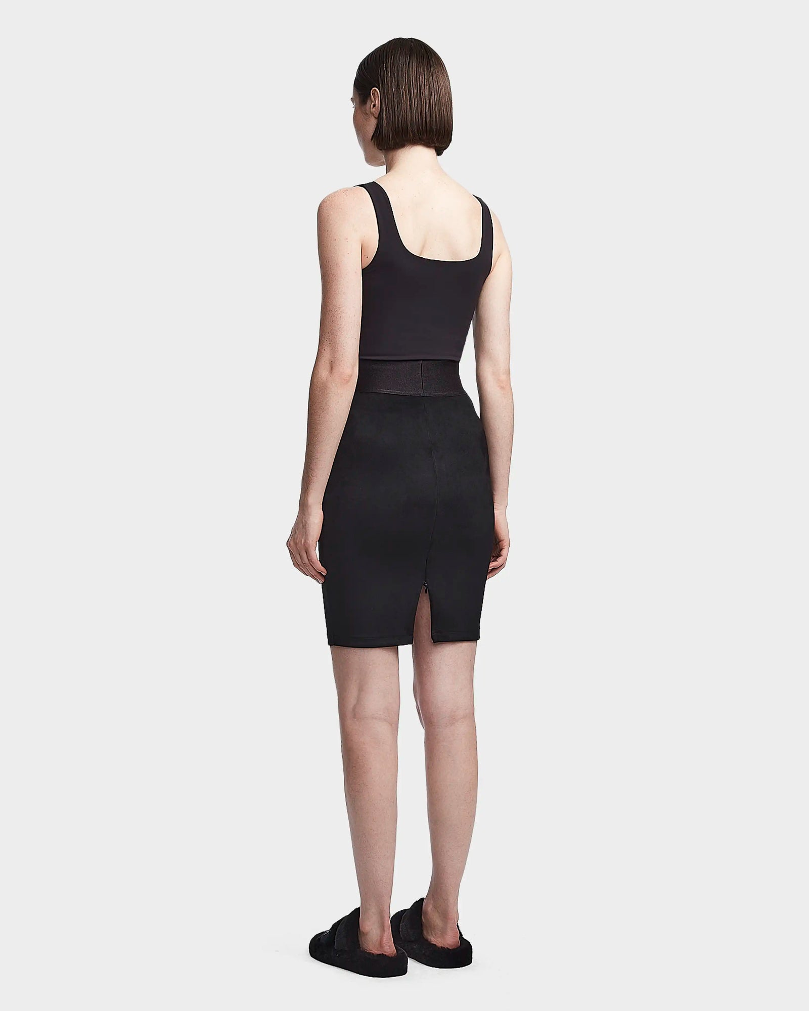 A woman with short brown hair faces away, wearing a sleeveless black top and the CHEYNA - 8523514 Black faux suede pencil skirt by Rudsak, paired with black slip-on sandals against a plain light gray background.