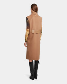 A person with straight brown hair models the Rudsak NOURA - 8124698 Camel knee-length vest over a printed long-sleeve top and knee-high black boots, standing with their back to the camera against a plain white background.