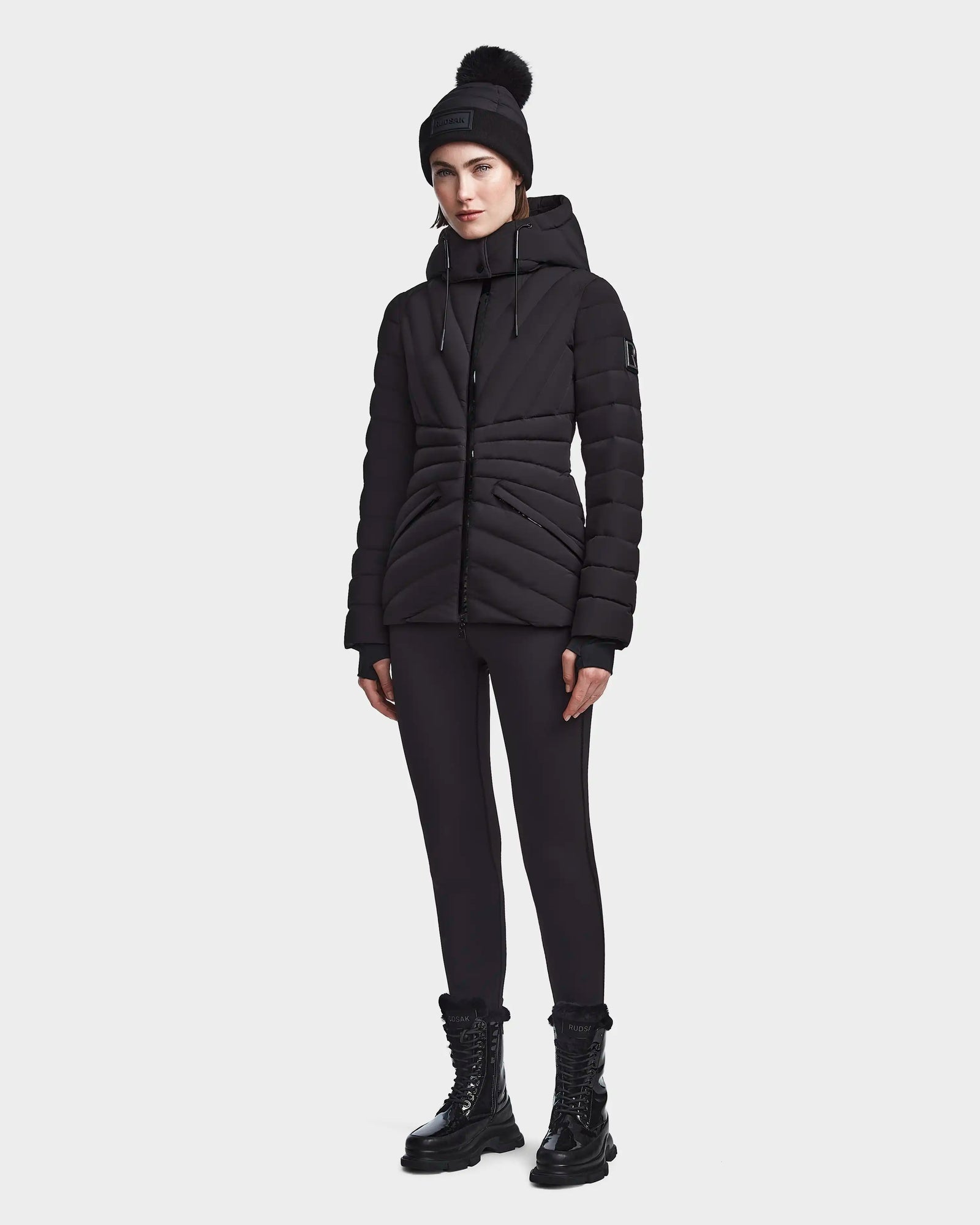 A person stands against a plain white background wearing the Rudsak HANNA - 8123562 Black down jacket, black leggings, black lace-up boots, and a black pom-pom beanie.