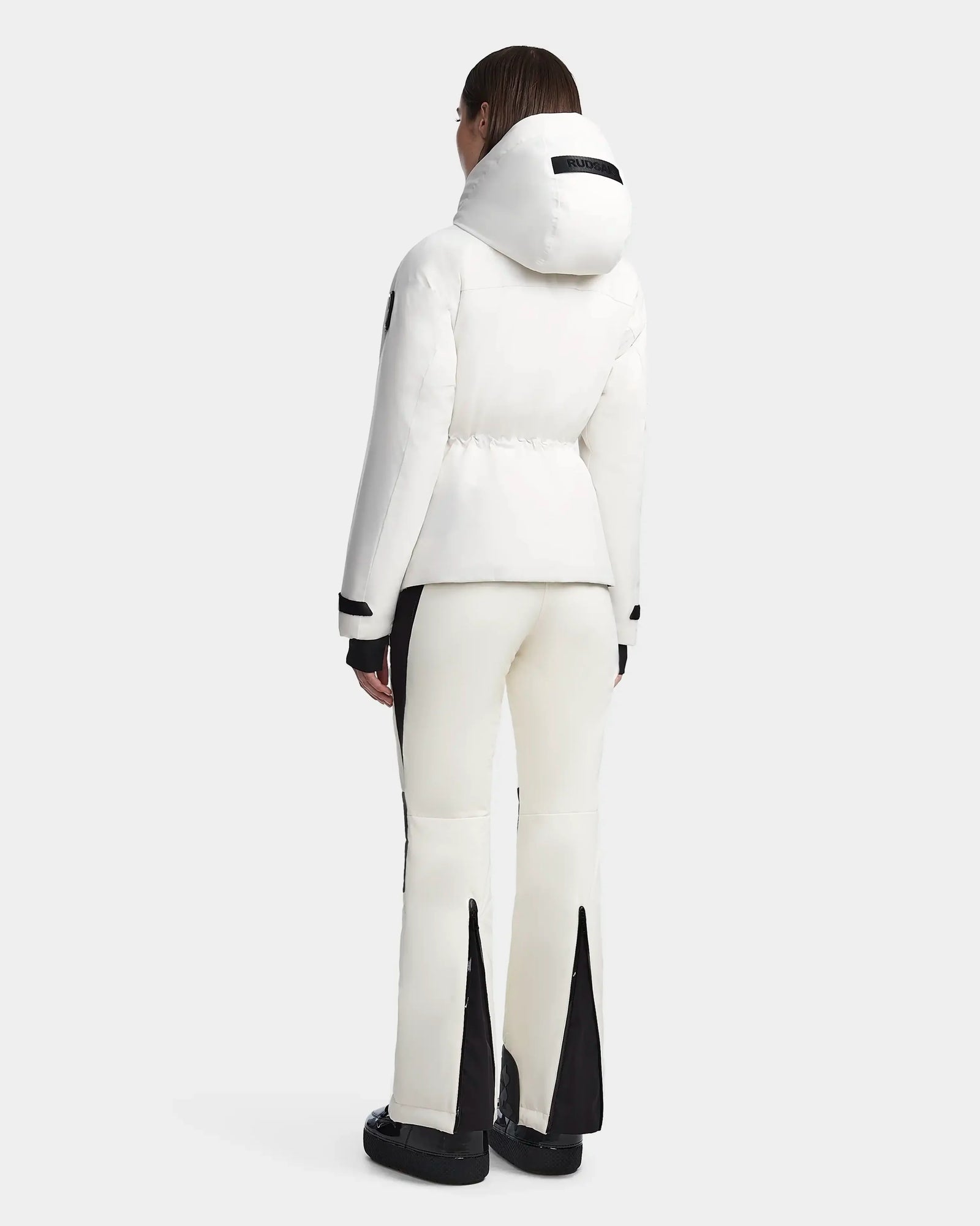 A person stands facing away, wearing the Rudsak LUX - 8123618 Iceland white waterproof ski jacket with RECCO technology and matching white pants featuring black panels and flared legs. They complete the sporty look with black shoes on a light gray background.