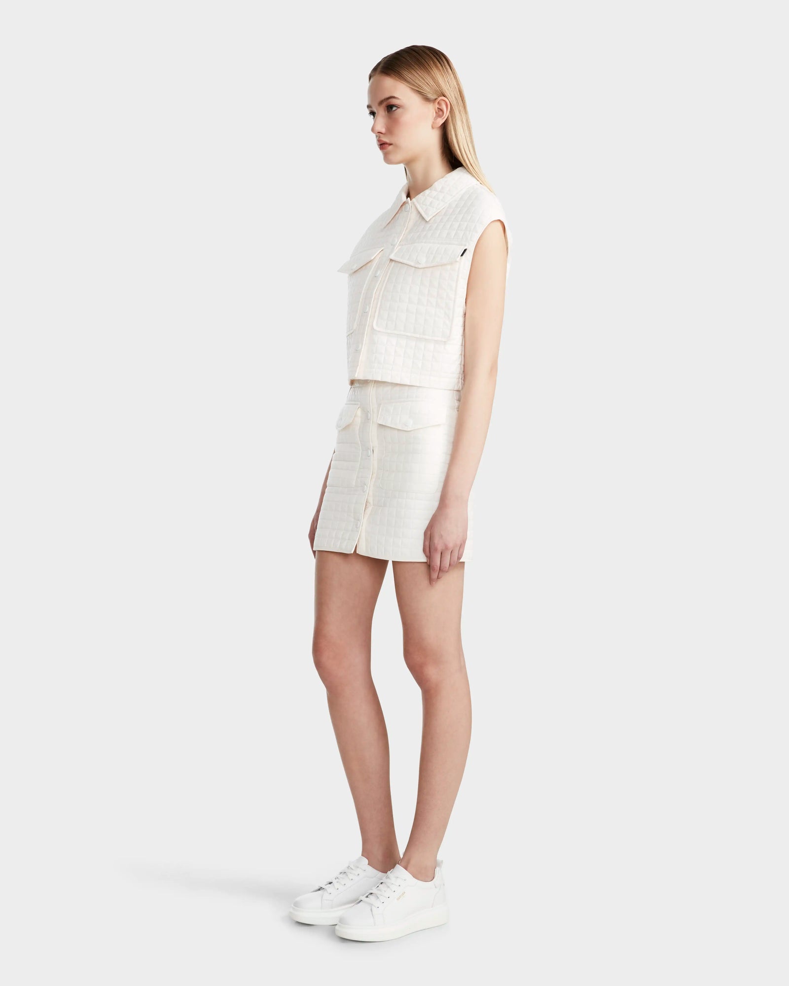 A woman with straight blonde hair stands sideways in the Rudsak SOLSA - 8124048 Eggshell womens cropped quilted vest with eco-friendly Sorona, layered over a sleeveless white top and matching skirt, paired with white sneakers against a plain background.