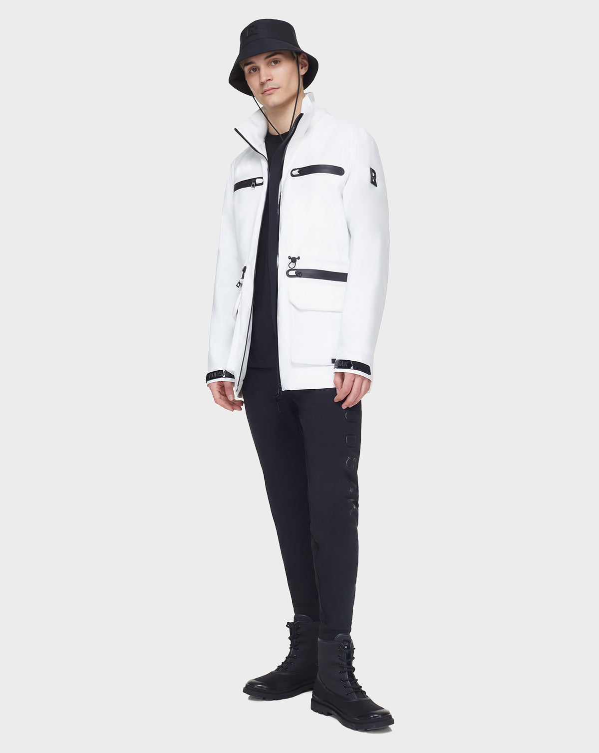 A person stands against a plain light gray background wearing the RUDSAK JACY - 6122000 WHITE mid-length raincoat, styled with black pants, black boots, and a black bucket hat.