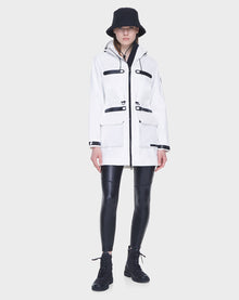 A person stands against a plain light gray background wearing the Rudsak CAMY - 8122002 WHITE water-resistant trench coat with black accents, paired with a black bucket hat, shiny black leggings, and lace-up boots.