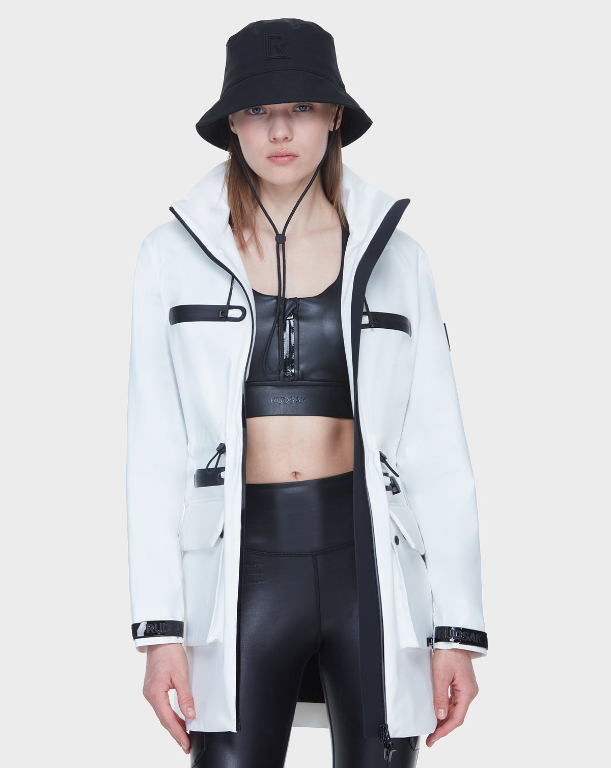 A person wearing the Rudsak CAMY - 8122002 WHITE mid-length raincoat with black zippers, a black bucket hat, a black crop top, and black high-waisted leggings stands facing forward against a plain white background.