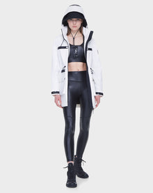 A woman faces forward wearing the Rudsak CAMY - 8122002 WHITE water-resistant trench coat over a black crop top, shiny black leggings, and black boots. The background is plain white.